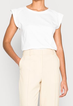 Top blanco de manga corta con cuello redondo y un ligero acolchado en los hombros, combinado con pantalones beige a medida. Tela suave y ajuste relajado.