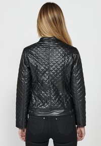 Giacca nera in finta pelle trapuntata con design aderente, polsini con zip e una texture liscia, caratterizzata da motivi a diamante e una vita.