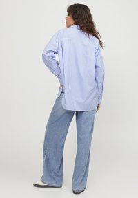 Camicia a righe blu e bianche, oversized, con maniche lunghe, polsini con bottone e orlo arrotondato, abbinata a jeans larghi azzurri.