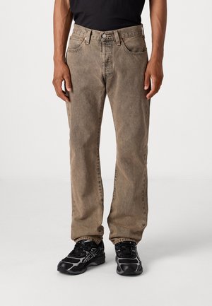 Homme portant un jean droit marron délavé et des chaussures de course noires, debout avec les bras détendus le long du corps, sur fond blanc.