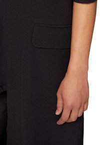 Černý oděv z tkaniny s viditelnou boční kapsou. Detail paže s rukou spočívající na materiálu, ukazující hladkou texturu.