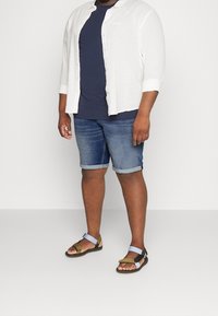 Bílá lněná košile s dlouhým rukávem, námořnické tričko, džíny s krátkými nohavicemi s ohrnutými manžetami a hnědé sandály s modrými akcenty. Neformální letní outfit.