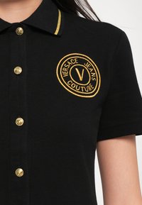 Čierne polo tričko vyrobené z textúrovanej látky, s pozlátenými gombíkmi a golierom so zlatým pruhom, a vyšitým okrúhlym logom s nápisom "Versace Jeans Couture."