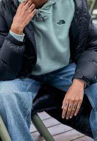 Giacca a vento nera sopra un hoodie verde chiaro, con coulisse e logo. Indossata con jeans a gamba larga di un blu chiaro. Tessuto testurizzato visibile.