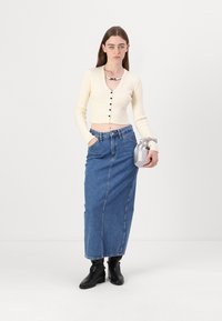 Krémový žebrovaný zkrácený kardigan s černými knoflíky, spárovaný s džínovou maxi sukní s vysokým pasem. Modelka drží malou stříbrnou tašku a má na sobě černé boty.