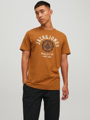 Brown cotton t-shirt featuring a round neckline, short sleeves, and a printed logo with a blue design. Worn with black pants.