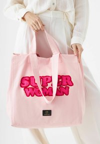 Sac tote en toile rose avec un texte brodé en peluche rose "SUPER WOMAN" et une étiquette noire avec "CODELLO" au bas.