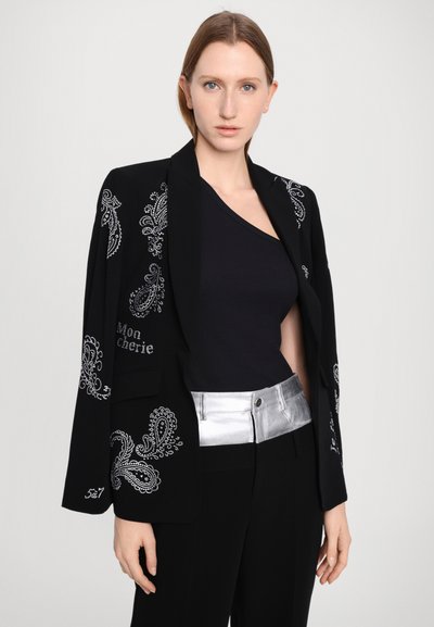 Zwarte blazer met zilveren paisley versieringen, ontwerp met één knoop, open voorkant, gedragen over een zwarte top, gecombineerd met zilveraccentueerde broek.
