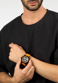 Orologio digitale con cassa nera, cinturino in silicone arancione e display trasparente. Indossato al polso, mette in risalto un forte contrasto di colori e dettagli.