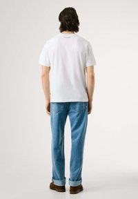 Mann med skulderlangt hår iført en hvit T-skjorte, lyseblå rullede jeans og brune sko, stående med ryggen mot kameraet på en ensfarget bakgrunn.