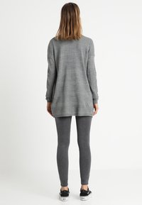 Lång grå tunika i texturerat stickat material, kombinerad med mörkgrå leggings och svarta sneakers, sedd bakifrån.