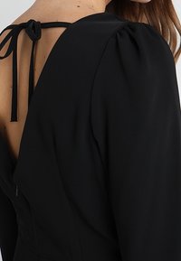 Vestido negro con espalda redondeada y detalle de lazo, hombros abullonados y una tela suave y texturizada. Cierre con cremallera en el lateral.