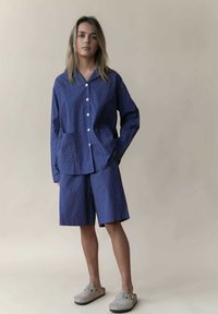 Marineblaues Polka-Dot-Hemd mit kurzen Ärmeln und passender Shorts, aus weichem Material, mit lockerer Passform und weißen Knöpfen. Graue Schlupfschuhen.