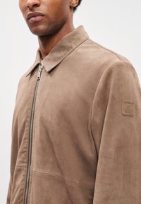 Barna velúr dzseki elöl cipzárral, hegyes gallérral és texturált felülettel. A bal vállon logófolttal díszített.