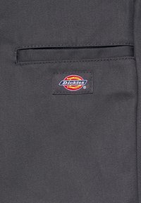 Tessuto grigio con una tasca a toppa e un'etichetta con il logo "Dickies" in rosso, giallo e blu, cucita sul materiale.