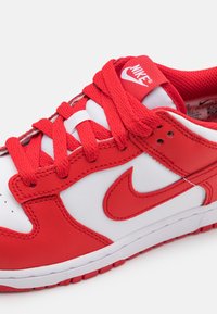 Raudoni ir balti Nike sportbačiai, turintys odinį viršų, tekstūruotą medžiaginę liežuvėlį ir plokščius raudonus raiščius. Išskirtinis Nike logotipas šone.