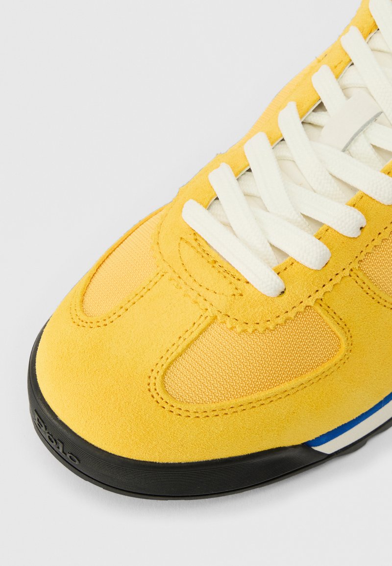 Gelbe Wildleder- und Mesh-Sneaker mit weißen Schnürsenkeln und schwarzer Sohle, die nahe der Ferse einen kleinen blauen und weißen Streifen aufweisen.