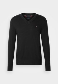 Pull noir à manches longues avec col en V et petit logo rouge, blanc et bleu sur le côté gauche de la poitrine, présenté sur fond blanc.