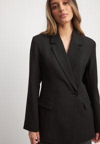 Melnā pielāgotā žakete ar izgrieztu uzaci un vienu pogu, kas lepojas ar divām priekšējām kabatām un strukturētu piegriezumu.