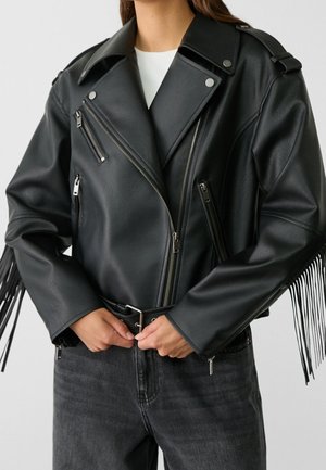 Persoon die een zwarte leren bikerjack draagt met zilveren ritsen, een taille met riem en franjes op de mouwen, gecombineerd met donkere jeans.
