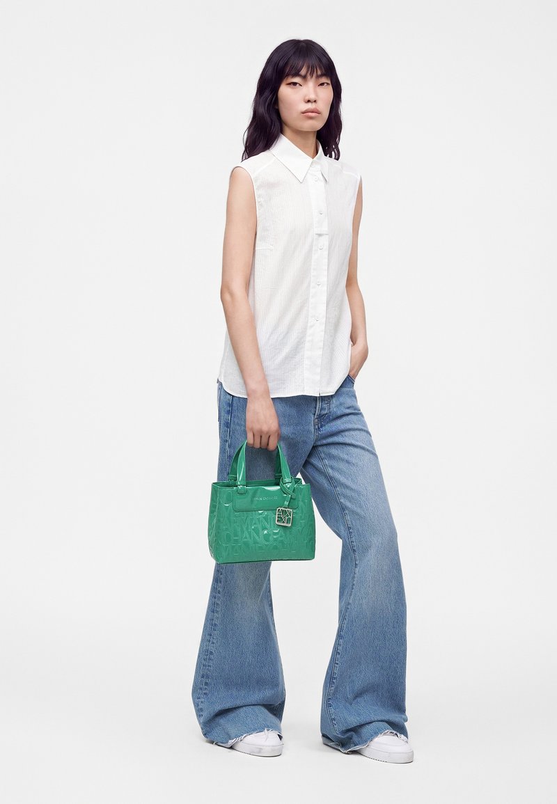 Jeune femme portant un débardeur blanc et un jean à jambes larges, tenant un petit sac à main vert sur fond blanc.
