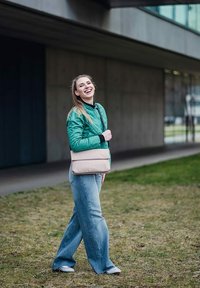 Zelená prošívaná bunda, černý top, široké světle modré džíny a pohodlné boty; nese béžovou kabelku přes rameno; stojí na trávě s neutrálním pozadím.