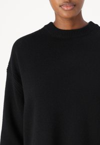 Črn pleten pulover z rebrastim krojem, spuštenimi rameni in mehkobo tkanine. Poglej bližje poudarja podrobnosti tkanine.