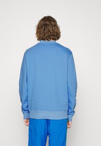 Personne avec des cheveux bruns bouclés portant un sweatshirt bleu uni et un pantalon bleu, se tenant de dos devant un fond blanc.