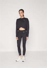 Schwarzes, kurzes langärmeliges Oberteil mit PUMA-Logo, kombiniert mit hoch geschnittenen schwarzen Leggings mit Seitennähten und hellen Sportschuhen.
