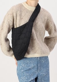 Schwarze gesteppte Schultertasche mit geschwungenem Design, einer strukturierten Oberfläche und einem breiten Riemen, getragen über einem gemusterten beigen Pullover und blauen Jeans.