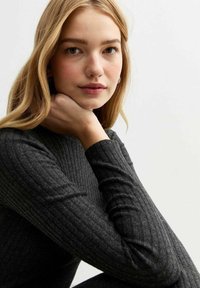 Geribbelde langzamige top in donkergrijs met een aansluitende pasvorm. Model steunt haar kin op haar hand en toont zachte texturen en subtiele details.