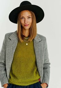 Šedý herringbone blazer přes olivově zelený svetr, doplněný širokým černým kloboukem. Dalšími detaily jsou vrstvené náhrdelníky a viditelný růžový akcent.