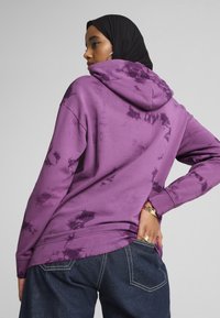 Ljubičasta hoodie s tie-dye uzorkom, s kapuljačom na vezanje i dugim rukavima, karakterizira opušteni kroj i suptilne varijacije uzorka. Nosena s tamnim trapericama.