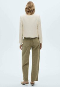Beige strukturierte kurze Jacke, lockere olivgrüne Hose und helle Schuhe, von hinten vor einem hellen Hintergrund gesehen.