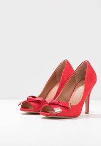 Escarpins rouges à talons hauts avec un dessus en tissu texturé, une conception à bout ouvert et des accents de nœud. Dotés de quincaillerie dorée sur le nœud.