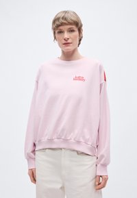 Svetloružová mikina s okrúhlym výstrihom, rebrovanými manžetami a lemom. Na hrudníku červené logo s textom "lolly's laundry". Mäkká textúra látky.