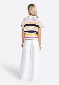 Haut en crochet avec des rayures multicolores en rose, jaune et noir. Manches courtes, tricot ajouré et ourlet en forme de vagues. Associé à un pantalon large blanc.