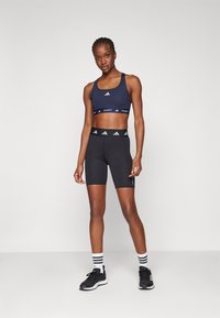 Marineblauwe sportbeha met wit logo en zwarte high-waisted shorts. Zwarte sportschoenen met witte strepen op zwarte sokken.