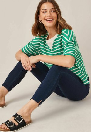 Tricou oversized cu dungi verzi și albe, blugi ajustați de culoare bleumarin cu fante la glezne și sandale negre cu două catarame, pe un fundal deschis.
