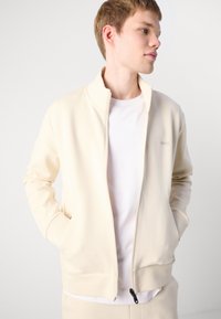 Beige rāvējslēdzēja jaka ar augstu apkakli, ribotām piedurknēm un divām sānu kabatām. Uz krūtīm ir maiga logo. Valkāta virs baltas krekla.