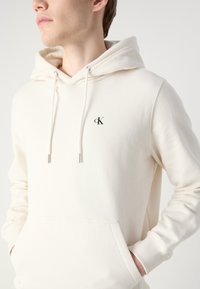 Sudadera con capucha de color beige claro, con bolsillo frontal, cordones y un pequeño logo negro en el lado izquierdo del pecho. Textura suave, diseño informal.