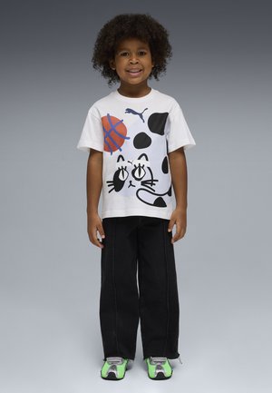 Niño con cabello rizado que lleva una camiseta blanca con un diseño de gato y baloncesto, pantalones negros y zapatillas verdes y grises, de pie y sonriendo.