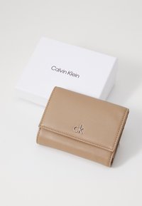 Béžová kožená peňaženka s preklápacím dizajnom, s embossovaným logom "ck" a striebornými akcentmi. Predstavená na bielej krabici Calvin Klein.