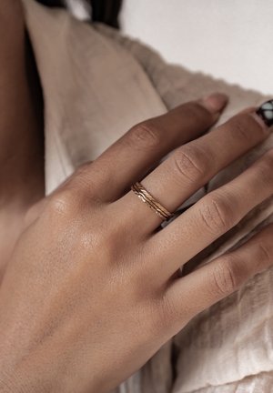 Anelli impilati in tonalità oro con design intrecciati e a spirale mostrati su una mano, caratterizzati da una pelle liscia e uno sfondo di tessuto testurizzato.