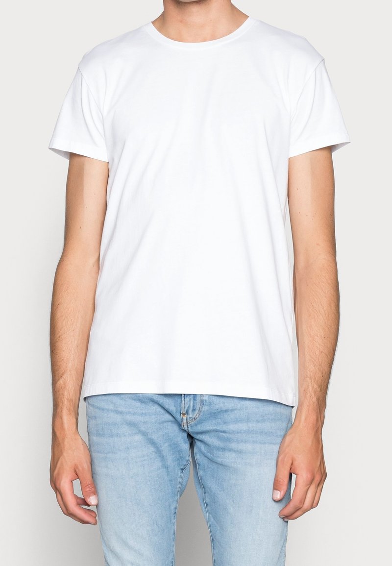 Homme portant un t-shirt blanc uni à manches courtes et un jean bleu clair, debout devant un fond neutre avec les bras détendus le long du corps.
