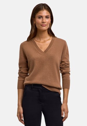 Brązowy sweter z dekoltem w serek, z długimi rękawami i prążkowanymi mankietami, wykonany z miękkiego materiału, w zestawieniu z ciemnymi spodniami. Prosty design bez wzorów.