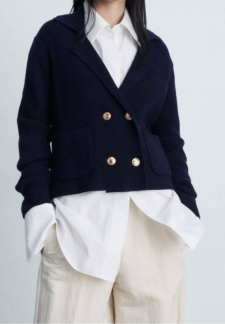 Femme portant un cardigan croisé bleu marine avec des boutons dorés sur une chemise blanche et un pantalon beige, mains dans les poches.