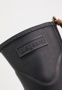 Botte de pluie en caoutchouc noir avec un bandeau supérieur texturé et un patch logo. Comprend une sangle en cuir brun avec des accents en métal.