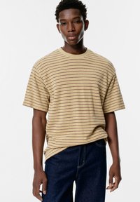 Tricou galben cu dungi, cu mâneci scurte și guler rotund, având linii subțiri roșii, purtat peste blugi negri.