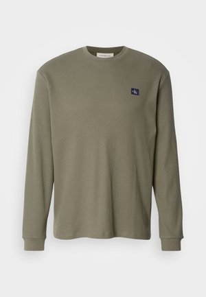 Pitkähihaisen oliivinvihreän ribbineulepaidan, jossa on pyöreä pääntie ja tummansinisellä logopatchilla vasemmassa rinnassa. Kangas näyttää olevan pehmeä ja teksturoitu.